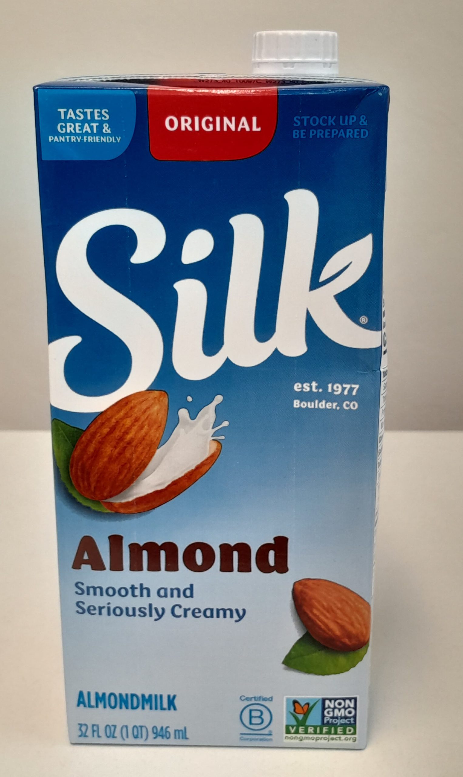 A front-facing photo of a 32-ounce carton of Silk Original Almond Milk. The blue carton features the Silk logo in large white letters, an image of creamy almond milk splashing out of a cracked almond, an almond in the lower corner, and labels indicating it is Non-GMO Project Verified and “Smooth and Seriously Creamy.'