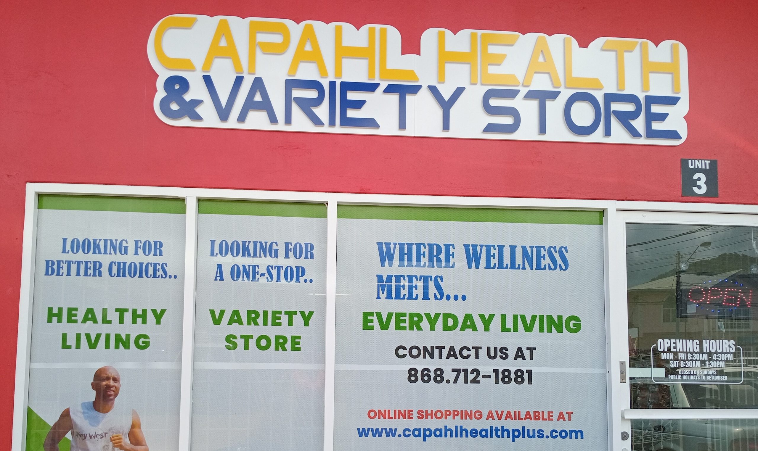 Storefront of CAPAHL Health & Variety Store with a red exterior and large white-and-blue sign. The front windows display printed graphics with text such as ‘Looking for better choices… Healthy Living,’ ‘Looking for a one-stop… Variety Store,’ and ‘Where wellness meets everyday living.’ A photo of a man jogging and images of herbs and spices appear on the window designs. The right side shows the glass entrance door with opening hours and an illuminated ‘OPEN’ sign.